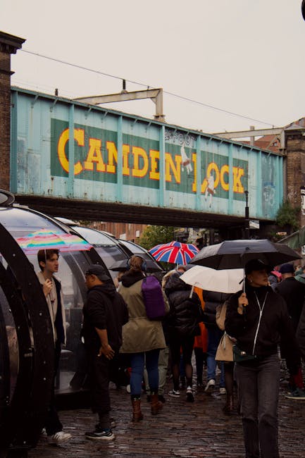 A crowded urban scene beneath a large, weathered blue railway bridge with the word 'CANDEN' partially visible, likely intended as 'CAMDEN' in yellow lettering. Pedestrians in various types of clothing