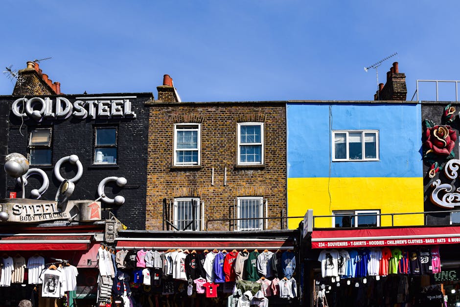 A crowded urban scene beneath a large, weathered blue railway bridge with the word 'CANDEN' partially visible, likely intended as 'CAMDEN' in yellow lettering. Pedestrians in various types of clothing
