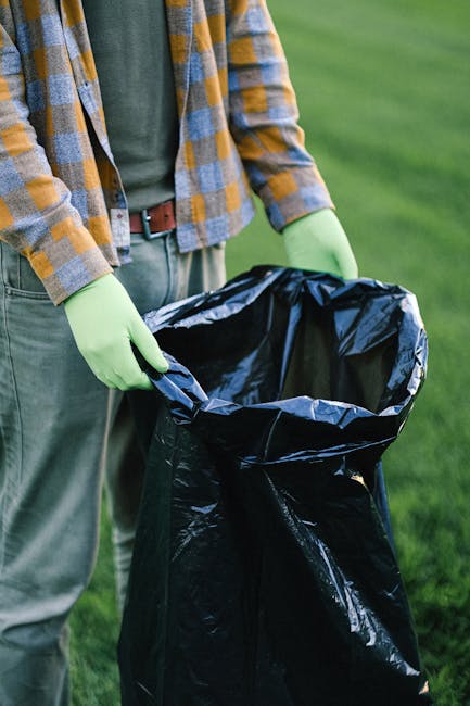 A waste collection worker wearing a high-visibility yellow and red vest is performing rubbish removal from the back of a large red garbage truck parked on the side of a street. The truck's rear compac