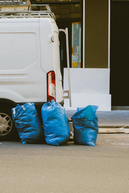 Close-up view of the rear side of a white commercial van parked on a street, with three large blue rubbish bags positioned on the pavement directly behind it. The bags appear to be filled with waste m